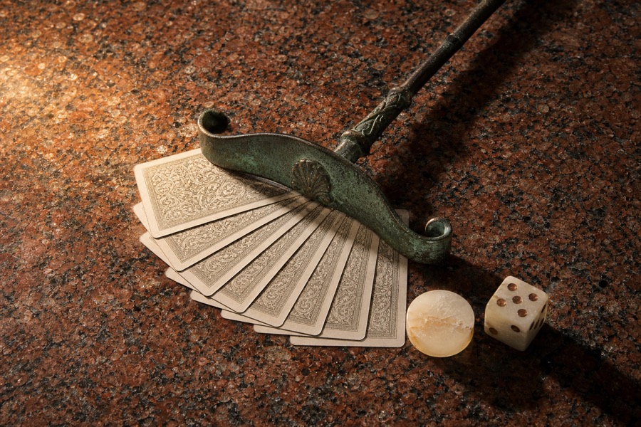 Rastrello da croupier in rame patinato posato su un ventaglio di carte da gioco coperte, con un dado e una fiche in alabastro sul granito rosso.